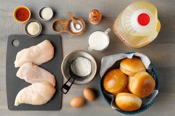 Top view of a kitchen counter displaying : raw chicken breasts on a cutting board, a bowl of flour, eggs, seasonings, hot sauce, milk, cooking oil, and a basket of brioche buns.